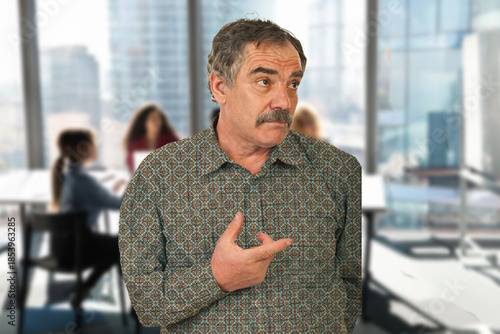 A man with a moustache pointing to his chest. He is in a room with other people