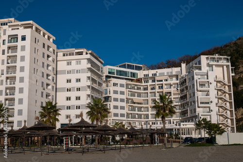A modern hotel by the sea and apartment buildings with palm trees and a sandy beach under a clear blue sky.