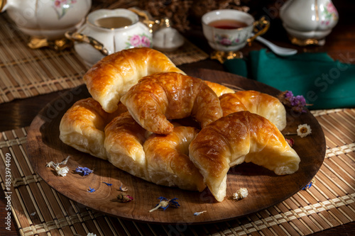 Plate with fresh sweet croissants for afternoon tea or breakfast, with a table set including tea and biscuits. 