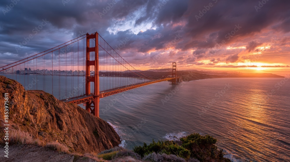 Fototapeta premium The Golden Gate Bridge connects San Francisco to Marin County as the sun sets. Clouds fill the sky while waves crash against the shore. This scene captures an evening in the city