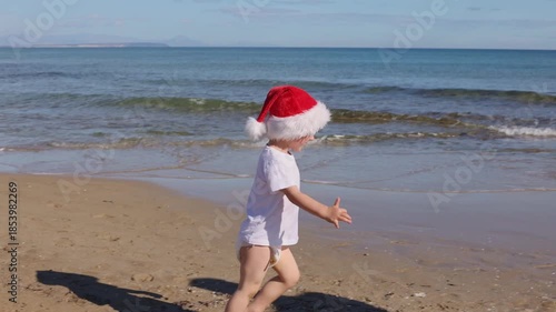 Happy little child wearing red Santa hat running and jumping on a sandy beach near sea. Christmas holidays concept. 
