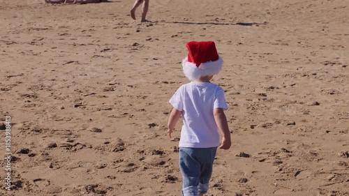 Happy little child wearing red Santa hat running and jumping on a sandy beach. Christmas holidays concept. 