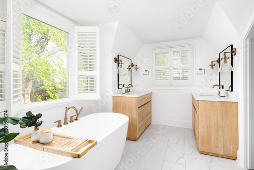 Bild auf Leinwand A beautiful bathroom with a freestanding soaking tub with a gold faucet, natural wood vanity cabinets, marble tile flooring, and bronze fixtures