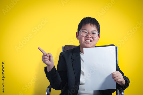 A boy in a suit sits in an executive chair, holding a white sheet of paper and pointing to a blank space against a gold background.