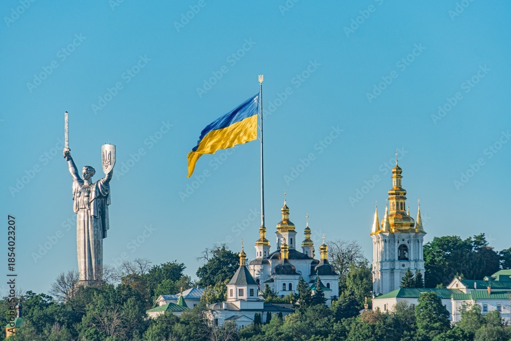 Obraz premium Panoramic view of Kyiv cityscape, clear sky, multiple domed cathedral, modern and traditional architecture, flag blue and yellow, statue