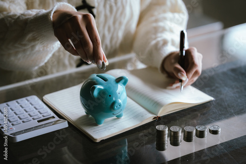 Blue piggy bank with stacked coins on a table, while a person uses a calculator in the background, representing saving money, budgeting, personal finance, and financial planning concepts.