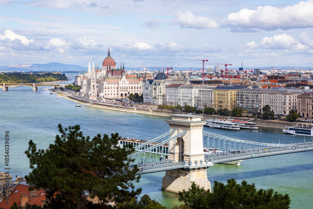 Obraz premium High angle panorama of trees, roofs and the 1849&nbsp;Széchenyi Chain Bridge over the Danube River and historic buildings including the Gothic Renaissance Parliament on the Pest side of the city, Budapest