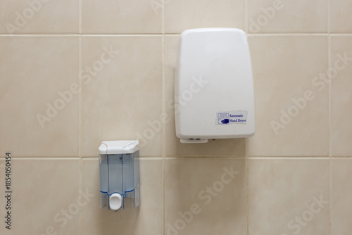 An air hand dryer hangs on the wall in the toilet, with a hand soap dispenser hanging nearby.