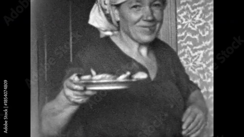 Smiling grandmother bringing home cooked food in rural home. Warm domestic holidays with elderly relatives. Everyday countryside life and traditional cooking. Old film. Archival retro. Vintage archive