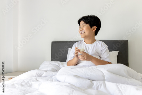 Asian boy holding glass of milk in bed at bedroom after waking up in morning. Sleepy chubby kid with milk glass in comfortable white bed. Portrait of Cute elementary school child drinking tasty milk.