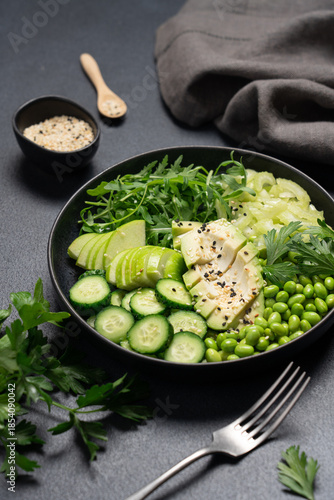 Healthy green salad with avocado, celery, apple, arugula, edamame beans, cucumber, and parsley in black bowl on dark background, top view