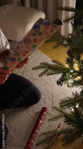 Unwrapping Christmas gift wrapping paper near decorated tree, holiday anticipation, festive New Year moment, hands opening present. 9:16