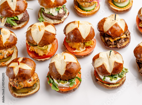 Close-up of mini burgers with fresh toppings. Mini burgers with pretzel buns, vegetables and cheese arranged on a white surface.