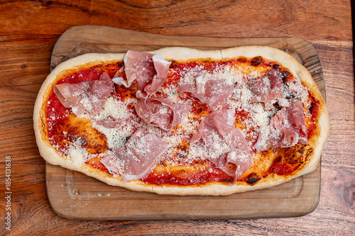 Italian pizza with prosciutto and parmesan on tomato base. Rectangular Italian pizza topped with tomato sauce, prosciutto slices and grated parmesan cheese on a wooden board.