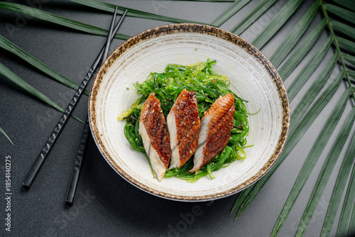 Grilled Eel Slices Over Fresh Green Seaweed Salad. Three pieces of perfectly glazed grilled eel rest atop a bed of bright green Japanese seaweed salad, served in a ceramic bowl.