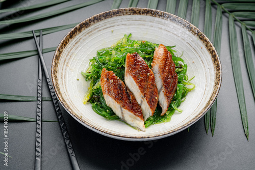 Grilled Eel Slices Over Fresh Green Seaweed Salad. Three pieces of perfectly glazed grilled eel rest atop a bed of bright green Japanese seaweed salad, served in a ceramic bowl.
