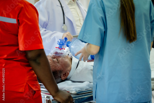 an elderly patient lying on a hospital stretcher receiving emergency medical treatment. Healthcare professionals in medical uniforms are using a manual resuscitator (Ambu bag) to provide assisted vent