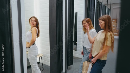 Woman stands outside fitting room showing new outfit to two friends who give feedback, concept of fashion choice and shopping experience