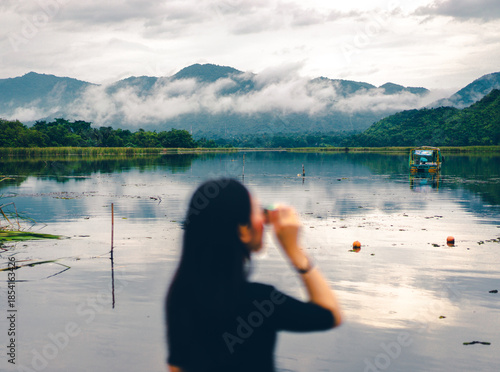Wallpaper Mural Blurred foreground of a woman standing with her back turned and holding a camera taking photos of landscape. A view of a mountain covered with fog and reflected in a pond in soft tone with copy space. Torontodigital.ca