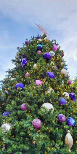 Close-up  of a large, lush Christmas tree adorned with numerous decorations and lights stretching all the way to the clouds in the winter sky; low angle shot, looking up the height of the tree