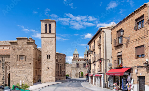 The Mudéjar tower of Santiago del Arrabal stands on the left, leading toward the Puerta de Bisagra gate in Toledo. Brick buildings and shops line the right side