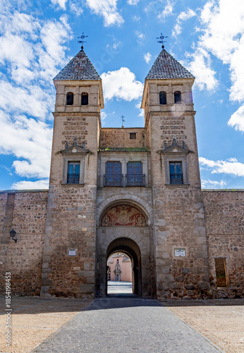 A grand view of the Puerta de Bisagra gate in Toledo, Spain, featuring two stone towers with tiled pyramidal spires and a central archway under a bright cloudy sky