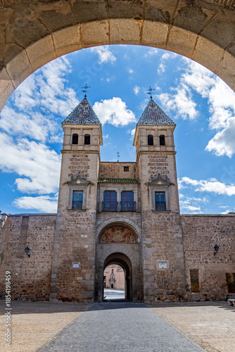 A grand view of the Puerta de Bisagra gate in Toledo, Spain, featuring two stone towers with tiled pyramidal spires and a central archway under a bright cloudy sky