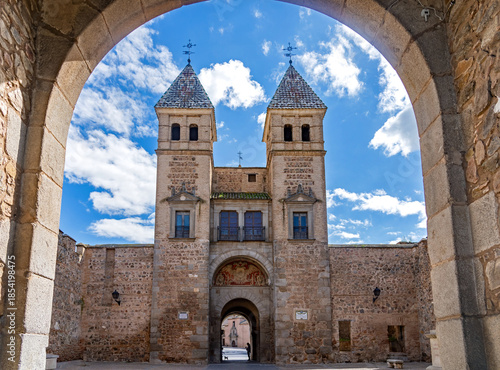 A grand view of the Puerta de Bisagra gate in Toledo, Spain, featuring two stone towers with tiled pyramidal spires and a central archway under a bright cloudy sky