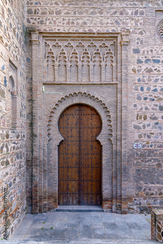 An intricate Mudéjar brick horseshoe arch frames a studded wooden door at Santiago del Arrabal in Toledo, Spain, set against ancient stone and decorative masonry