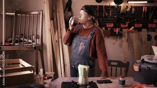 Female carpenter enjoying coffee break standing near workbench in modern woodworking repair workshop