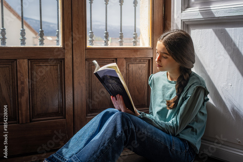 Preteen girl sitting by the window reading a book in natural light. Peaceful indoor moment portraying childhood, focus, and quiet learning at home.