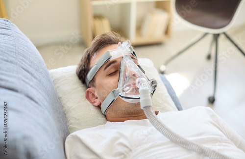 Man in aspirator with oxygen mask resting in bed. The patient gets breathing support during therapy for recovery. Clean setup suggests hospital healthcare at home. Calm care concept of recovery.