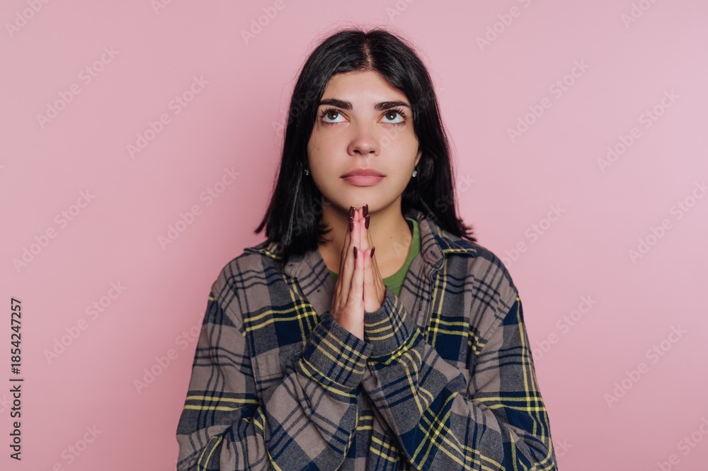 Fototapeta premium Young woman with hands together looking up against a pink background in a casual outfit during indoor time