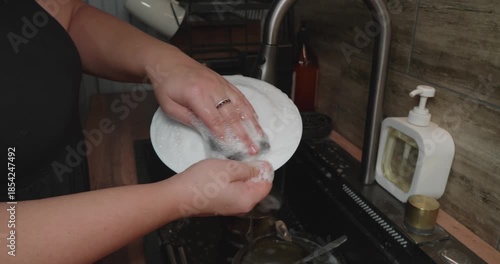 A close-up of a woman's hands carefully washing a white plate with a soapy sponge in a modern kitchen sink The woman rinses the plate under clean running water from the tap during her evening chores.