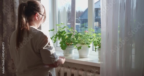 A diligent woman wearing glasses removes potted plants from a windowsill in a sunlit room before beginning spring cleaning and housework. The woman prepares the window for washing.