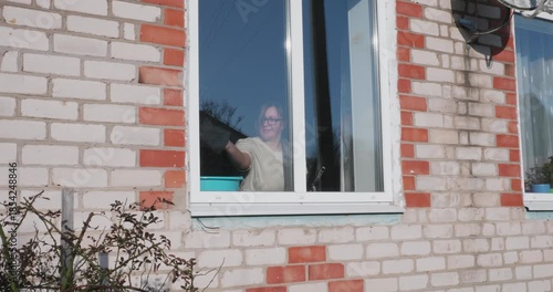A hardworking woman washes a large window from the inside of a brick residential building using a squeegee, smiling as she goes about her daily chores. The woman is washing windows in a house.