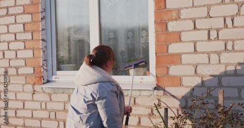A hardworking woman in a jacket washes the outside window of a brick house with a long brush while doing chores in the courtyard on a sunny day. The woman is washing a window facing the street.