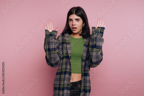 Woman holds hands up in surprise wearing green top and checkered jacket against a pink background in indoor setting