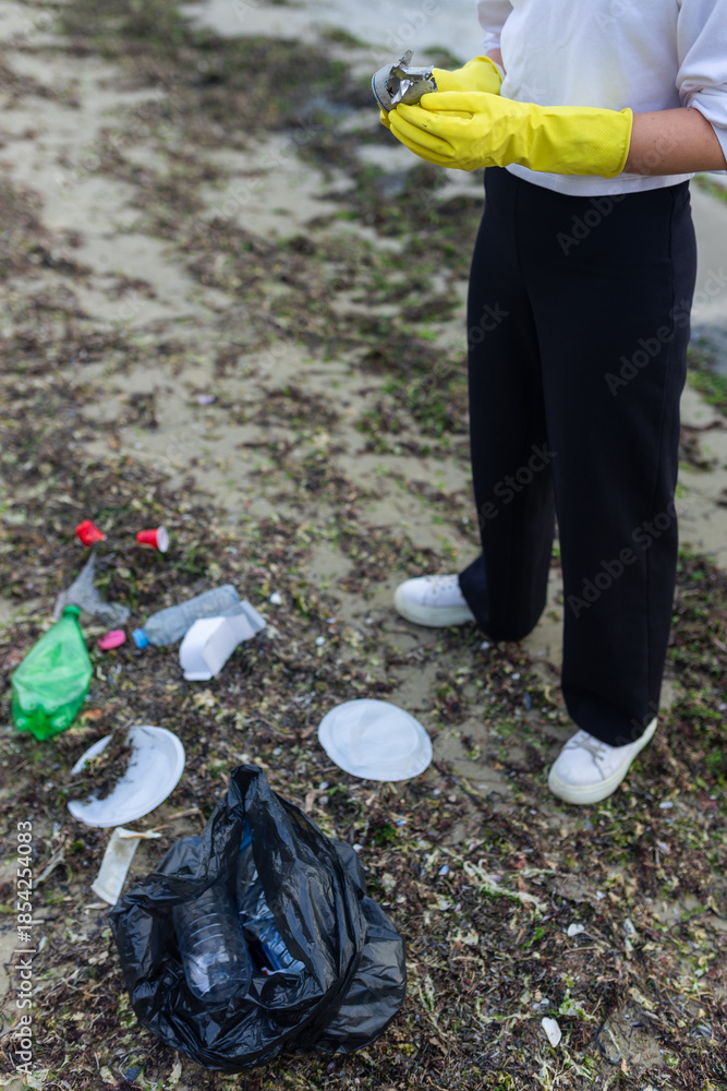 Obraz premium volunteer cleaning polluted beach, collecting plastic bottles, cups and debris in a black bag, supporting ecology, environment protection, sustainability and coastal cleanup awareness.