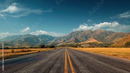 A road runs straight through a wide valley with mountains in the background. The sun shines brightly. Grass and trees line the sides of the road.