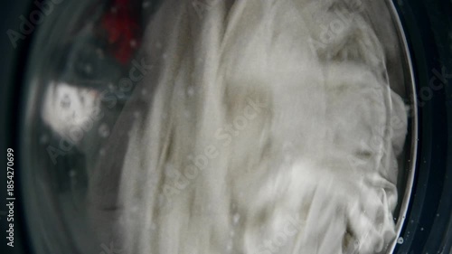 A close-up of old, yellowed gray laundry that was once white being washed in a washing machine. Bubbles of detergent drip down the glass window.