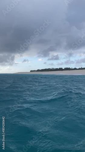 Vertical POV video of deep blue ocean waves moving rhythmically towards a small, uninhabited tropical island under a cloudy sky, emphasizing travel and natural beauty.