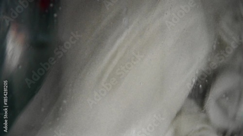 A close-up of old, yellowed gray laundry that was once white being washed in a washing machine. Bubbles of detergent drip down the glass window.
