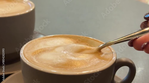 A close-up of a glass cup filled with a hot coffee latte, topped with thick, creamy foam, and a spoon resting inside, perfect for coffee lovers