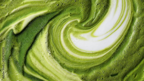 A mixture of matcha and milk is swirling together in a bowl. The colors blend to create a gradient of green and white. This shows the process of making a drink at home in the morning.