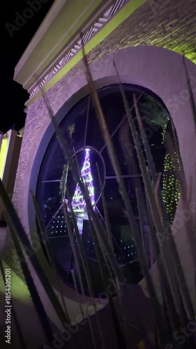 An illuminated Christmas tree with garlands is reflected in a round window at night