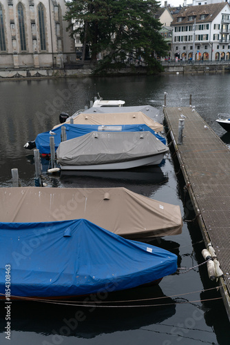 A calm European riverside cityscape with historic architecture, a church tower, and boats moored at a marina under an overcast sky.
