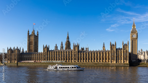 Obraz na płótnie Wide view of the Palace of Westminster and Big Ben along the River Thames in London, featuring historic Gothic architecture, a passing river boat, and clear blue sky on a sunny day