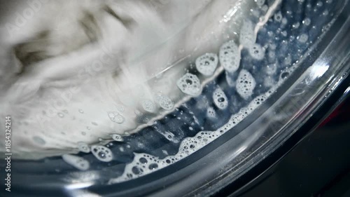 A close-up of old, yellowed gray laundry that was once white being washed in a washing machine. Bubbles of detergent drip down the glass window.