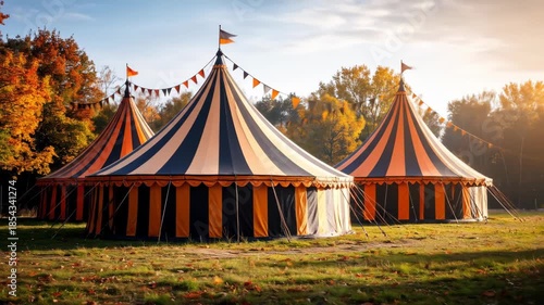 Three circus tents with orange and black stripes in a grassy field with autumn foliage.
